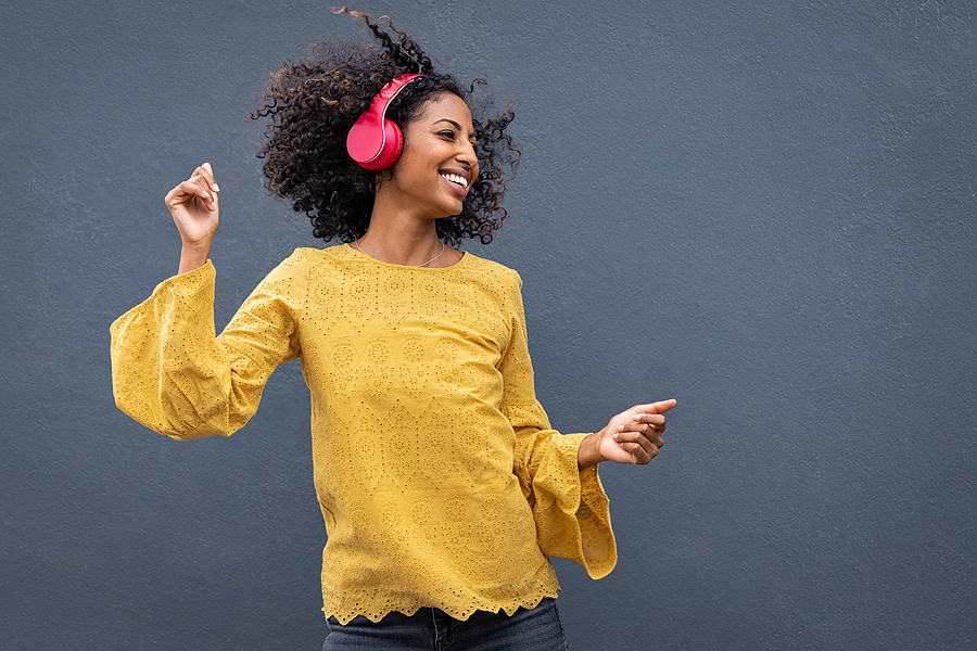 5 Amazing Portable Bluetooth Speakers You Would Like to Own Beautiful black girl listening music. Cheerful young woman with curly hair making dance moves to the rhythm of music while listening through wireless headphones isolated on grey wall.