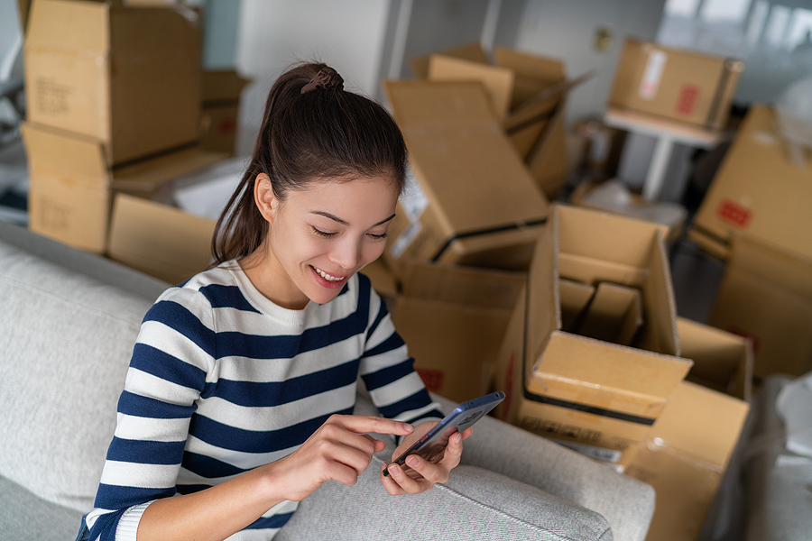 From Renting to Owning: A Beginner's Guide to Buying Your First Home Homeowner Asian woman using cellphone mobile phone for indoor mess with many open packages moving boxes for move in new apartment. Happy girl in condo house.