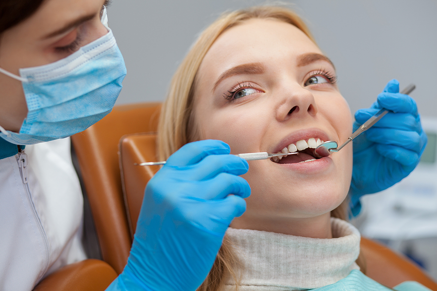 Attractive female dentist adjusting dental lamp, preparing to examine teeth of her patient. Professional dentist working at her clinic. Woman visiting dentist for teeth examination. Profession concept