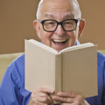 7 Mental Health Benefits of Reading Books Every Day Laughing senior male reading a book on a couch. Vertical