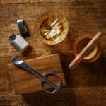 View of a crystal glass of golden whisky, cigar and cigar cutter and petrol lighter, shot from above on a dark wooden background