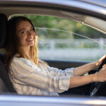 Happy young girl sitting on driver seat in new car joyful smiling hold hands on wheel. Cheerful female driving vehicle looking through open window. Successful woman car owner or getting driver license