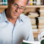 Mature man reading a book with glasses in a library. Looking at camera with confidence.
