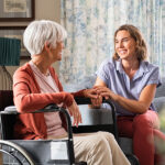 Mature woman comforting senior mom sitting on wheelchair at nursing home. Cheerful woman talking to old disabled mother at elder care centre. Loving caregiver taking care of elderly woman at home.