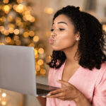 How to create the perfect online dating profile? Virtual Or Long Distance Relationship And Online Dating. Portrait of beautiful young curly black woman holding laptop computer in hands and sending kisses to web camera during video chat with partner