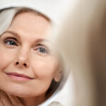 Vaginal Rejuvenation at Home Happy 50s middle aged woman model touching face skin looking in mirror reflection. Smiling mature old lady pampering, healthy moisturized skin care, aging beauty, skincare treatment cosmetics concept.