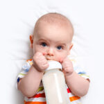 Caucasian Baby Boy With Baby Milk Bottle On White Blanket. Close