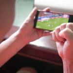 Sports fan with fist up in celebration while watching sports streaming on mobile phone. Close up view.