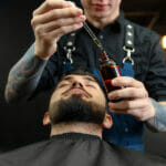 How to get the best results when using beard oil? Hairdresser With Beard Oil Near Client In Barbershop, Closeup. P