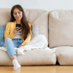 Is "Sliding into Instagram DMs" Still a Thing to Find Dates in 2025? Smiling millennial woman relax sit on couch at home, using phone, texting chatting with friend. Happy girl resting on sofa hold smartphone, shopping online, check mobile application or social media.