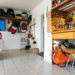 What Should Not Be Stored In A Garage? Home suburban car garage interior with wooden shelf, tools equipment stuff storage warehouse on white wall indoor. Vehicle parked at house parking background. DIY workbench for repair home appliances