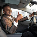 Cheerful smiling African man businessman, sitting in his new high tech electric car with upgraded avtopilot self-driving system, holding take away coffee and showing thumb up