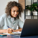 Willpower or Smart Time Management: How to Become a Successful Student Young African American girl student studying virtual course, learning distance class web training, looking at laptop computer watching online webinar and writing notes sitting at home office table.