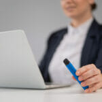 Buying a Disposable Vape? Here Are the 4 Reasons Why You Shouldn’t Business woman holding disposable vape while sitting at laptop.