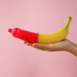 Woman holding banana in condom on pink background, closeup. Safe sex concept