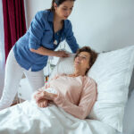 Home Nurse Making Elderly Patient Bed. Female Caregiver Adjustin